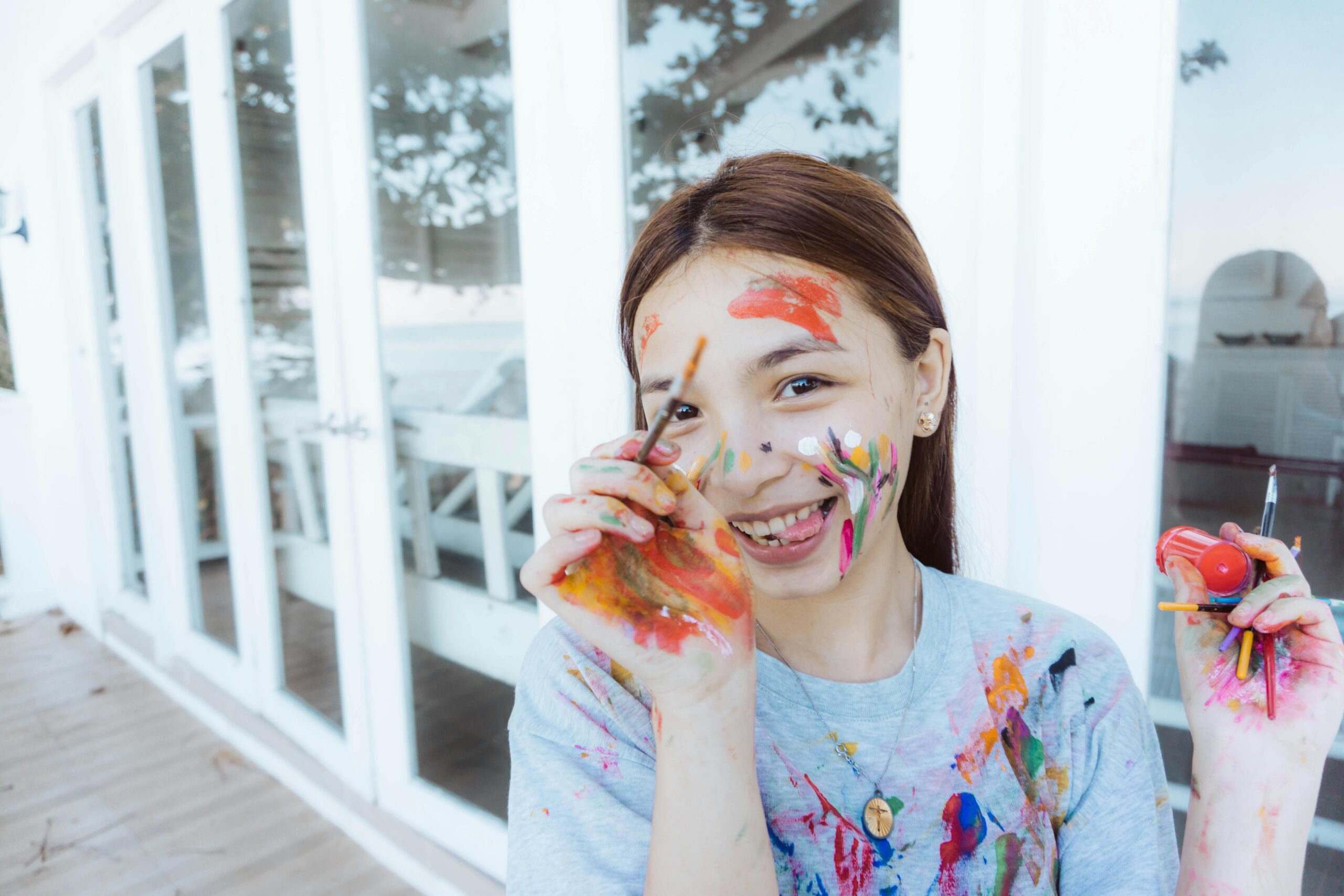 A cheerful woman covered in colorful paint holds brushes, showcasing her artistic creativity.